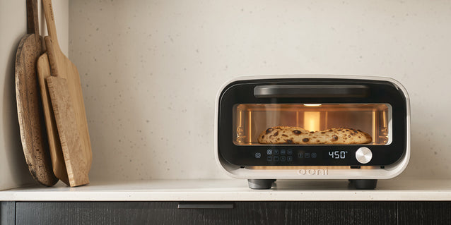 Ooni Volt 2 with pizza inside on a kitchen shelf next to wooden paddles.