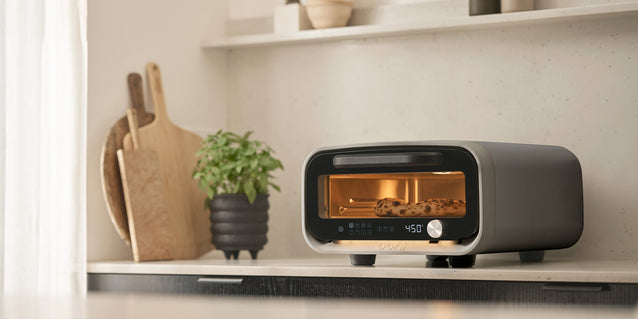 Ooni Volt 2 pizza oven on a kitchen shelf next to a plant and cutting board.
