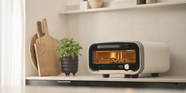 Ooni Volt 2 pizza oven on a kitchen shelf next to a plant and cutting board.