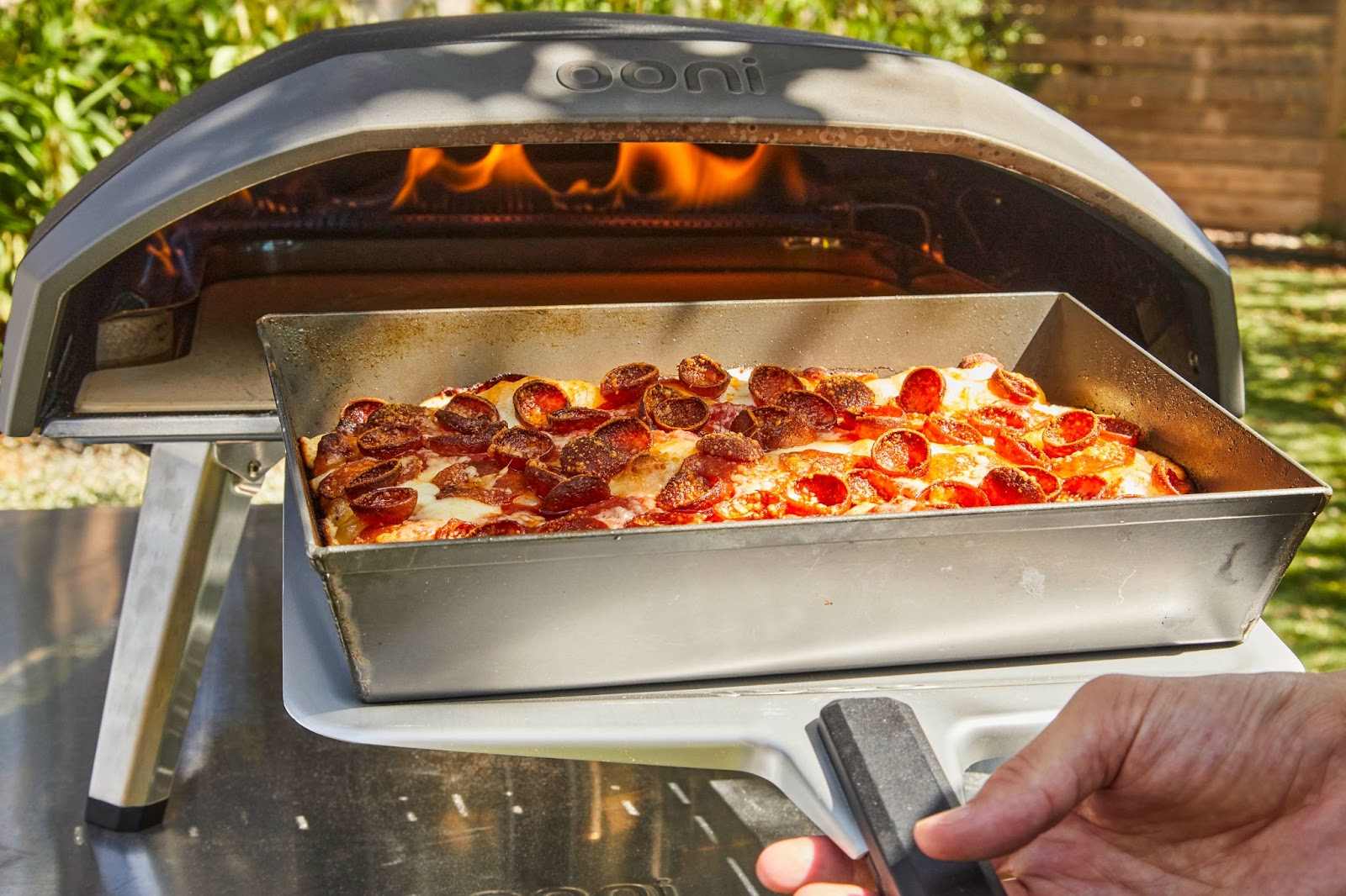 Hand holding an Ooni Pizza Peel with an overloaded pepperoni pizza in a deep dish pan in front of an Ooni oven with flames.