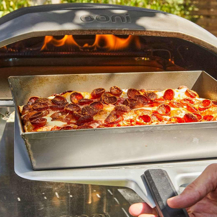 Hand holding an Ooni Pizza Peel with an overloaded pepperoni pizza in a deep dish pan in front of an Ooni oven with flames.
