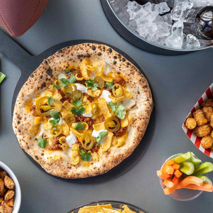 A pizza topped with fritos on a tabletop surrounded by snacks