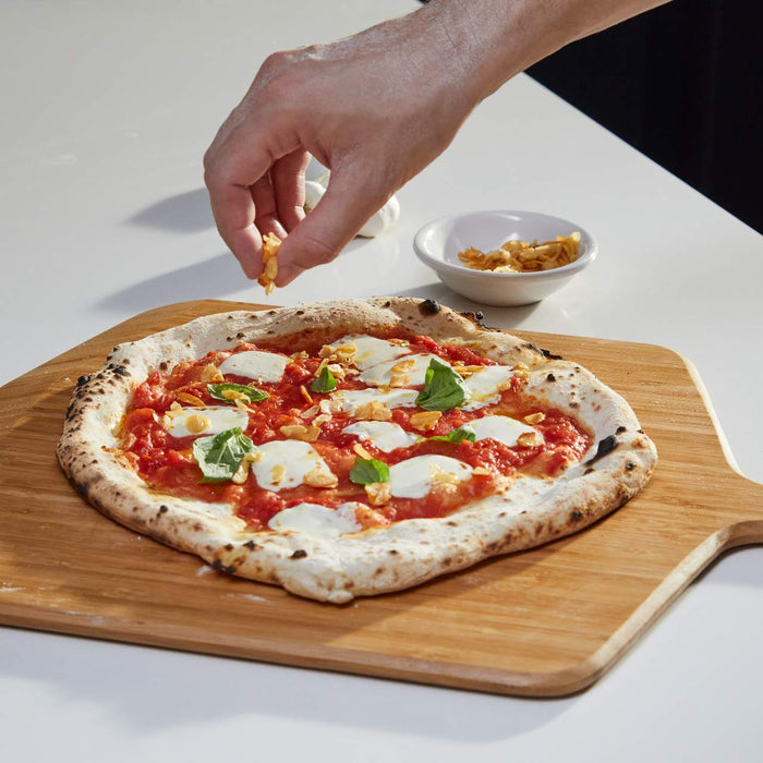 Pizza topped with cheese, tomatoes, basil being sprinkled with fried garlic chips on a wooden pizza peel