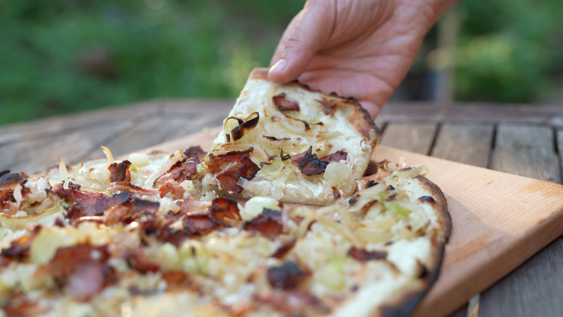 Hand taking a slice of cooked flammkuchen with crème fraîche, bacon lardons and onions on a bamboo serving board.