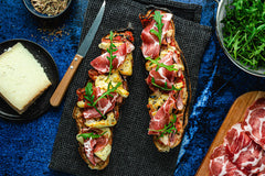 Cooked Corsican pizza baguette with Corsican cheese, pancetta, coppa, rocket and herbes de Provence on a table surrounded by a plate of cheese, bowl of rocket and serving board of pancetta.