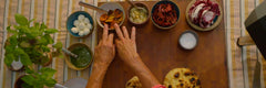 Two hands touching a bowl of marinated baby bell peppers next to bowls of basil pesto, mozzarella balls, garlic confit, Cruschi peppers, radicchio and salt.