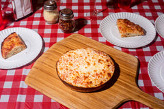 Cooked pan pizza with tomato sauce and mozzarella on an Ooni Bamboo Peel & Serving Board on a table with plates of slices of pan pizza.