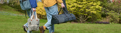 Two people walking through a park carrying an Ooni pizza oven in a black oven cover and a checkered tote bag with a wooden utensil handle marked ooni.