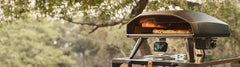 The Ooni Koda 2 Max on a metal table outdoors with trees in the background 