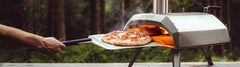 A person removes a cooked pizza from the Karu 12 oven with a blurred forest background