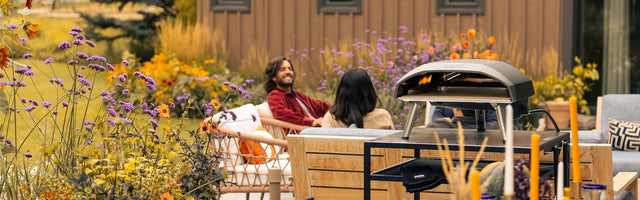 An Ooni pizza oven sits on a modular table in a vibrant garden, with the logo 'ooni' visible on the oven and a nearby glove. In the background, friends relax in a blooming outdoor setting.