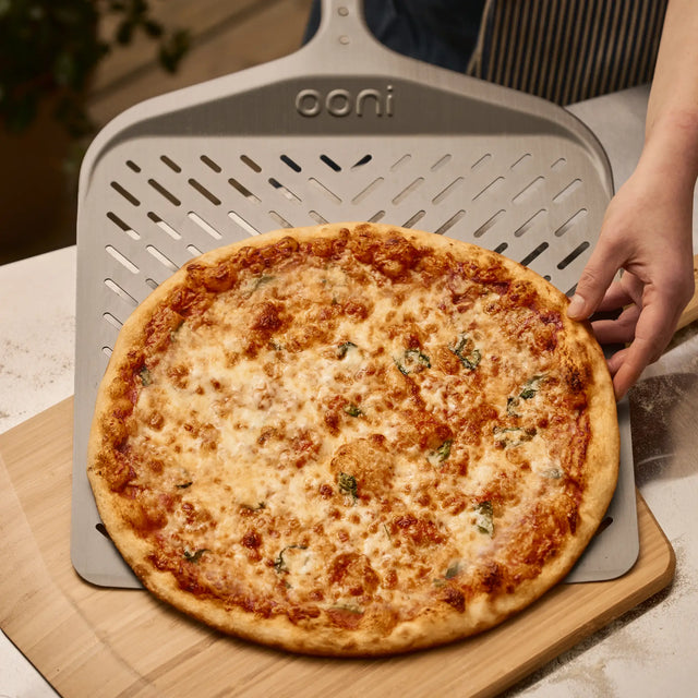 Person sliding a freshly baked pizza off the Signature Pizza Peel onto a bamboo pizza peel on a table.