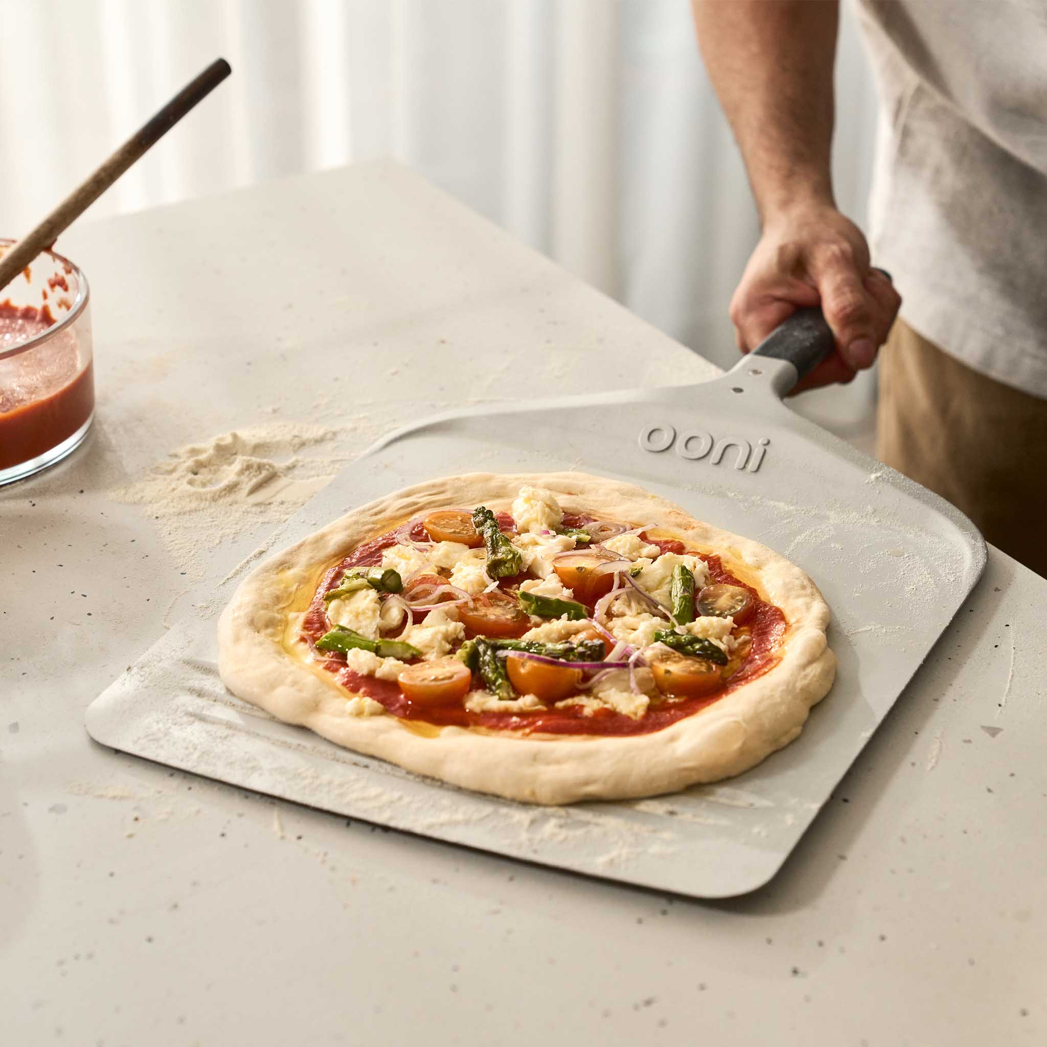 Prepped pizza on Ooni Compact peel with semolina & tomato sauce on kitchen countertop.