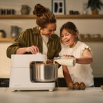 A woman and a young girl are baking together in a warm kitchen setting. The woman is wearing an olive green shirt, and the girl is in a white ruffled top.