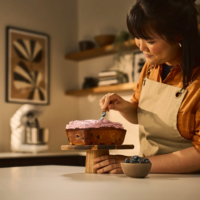 A woman in a kitchen wearing an apron and rust-colored shirt spreads pink frosting onto a loaf cake on a wooden stand. A small bowl of blueberries sits on the counter nearby, with shelves and framed wall art blurred in the background.