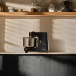 A black Ooni Halo spiral mixer with a stainless steel bowl sits on a kitchen counter, with warm sunlight casting windowpane shadows on the background wall and wooden shelf.