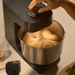 A black Halo Core mixer with a stainless steel bowl containing dough, with a hand adjusting the top dial. A small bowl and a black spatula are visible on a light beige surface.
