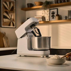A white Ooni Halo Pro mixer with a stainless steel bowl sits on a kitchen counter, with a bowl of flour in the foreground and a softly blurred kitchen interior behind it.