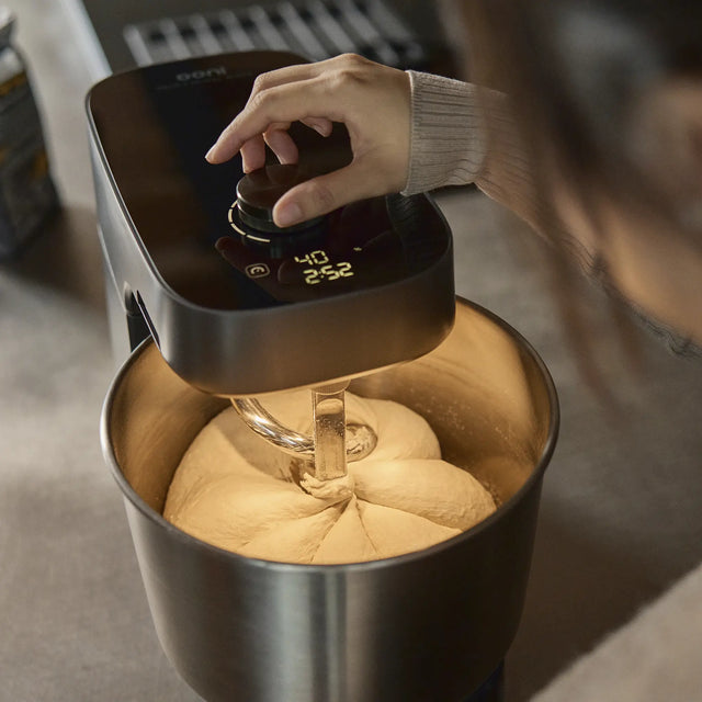 A hand adjusts the control dial of the Halo Pro stand mixer with a stainless steel bowl containing dough, set against a softly blurred kitchen background.
