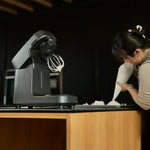 A Halo Pro mixer with a whisk attachment sits on a wooden counter next to a woman piping white cream from a bag.