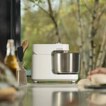 A white Halo Pro with a stainless steel bowl sits on a reflective countertop in a kitchen setting, with a blurred window showing trees in the background and dough visible on the right edge.