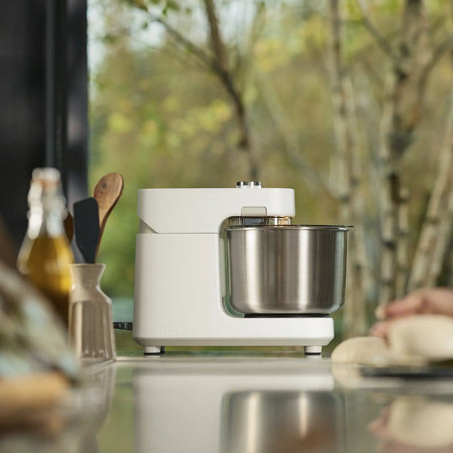 A white Halo Pro with a stainless steel bowl sits on a reflective countertop in a kitchen setting, with a blurred window showing trees in the background and dough visible on the right edge.