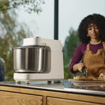 A white Halo Pro mixer with a stainless steel bowl sits on a kitchen counter, with a woman in an apron preparing food in the background.