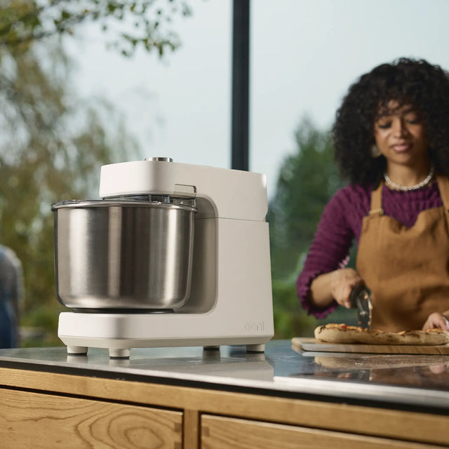 A white Halo Pro mixer with a stainless steel bowl sits on a kitchen counter, with a woman in an apron preparing food in the background.