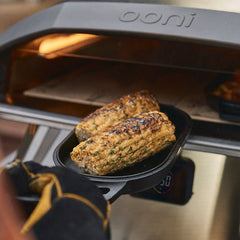A pair of gloved hands holding a cast iron sizzler pan with grilled corn 