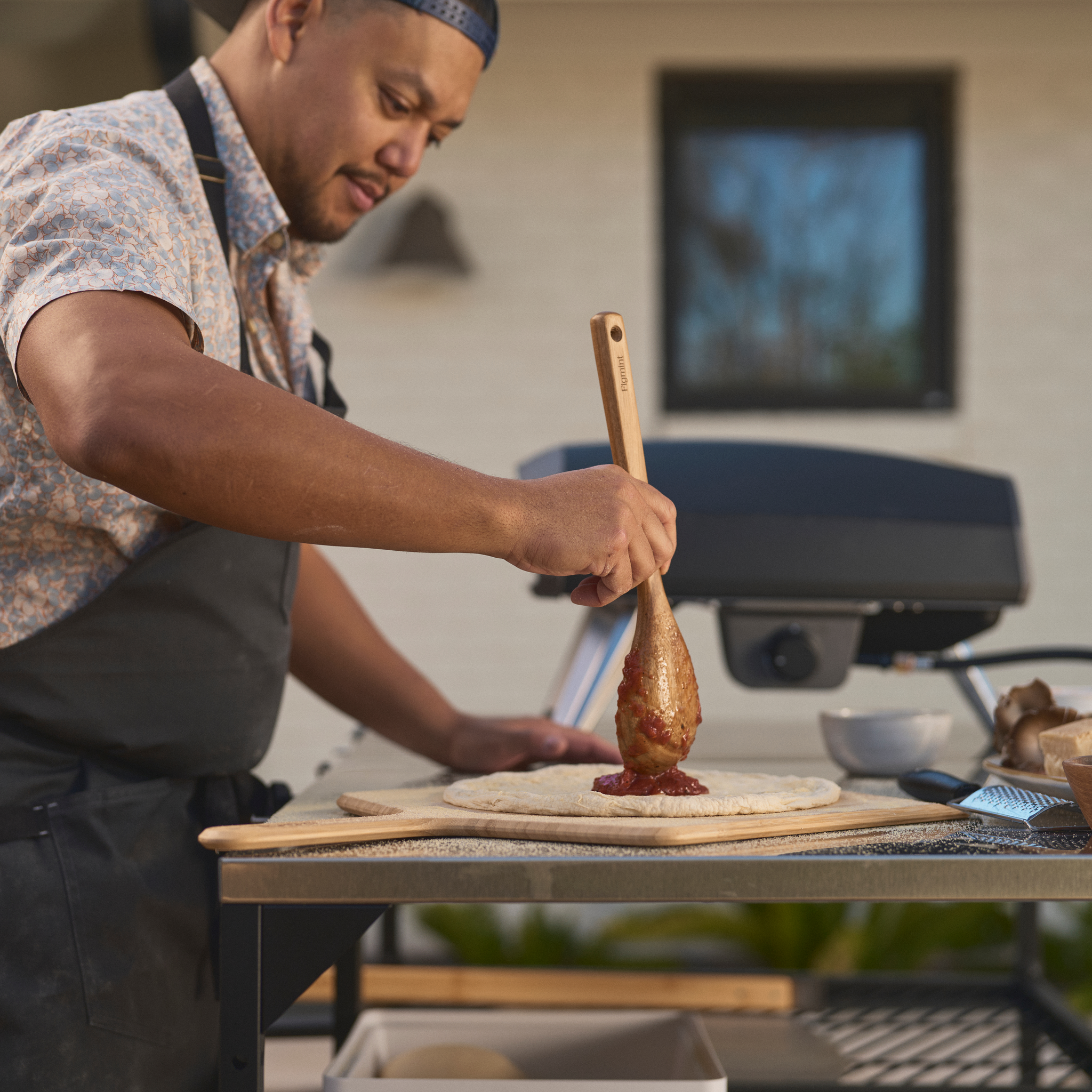 Person preparing pizza with a wooden spoon in an outdoor kitchen setting with the Ooni Koda 2 Gas Powered Pizza Oven in the background