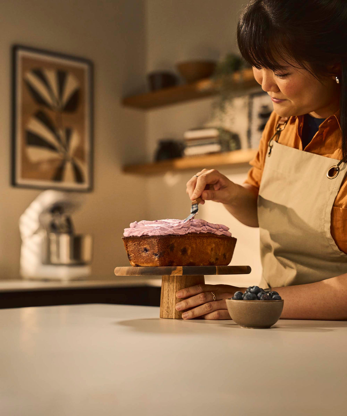 A woman icing a loaf cake with a blurred kitchen background