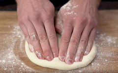 Flour-dusted hands stretching a fresh pizza dough ball on a wooden prep surface.
