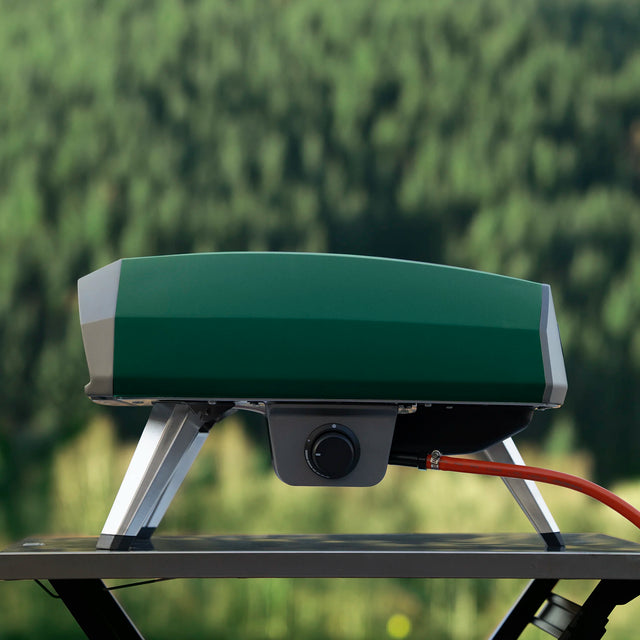 The Highland Green Koda 2 pizza oven on a camping table with a blurred green forest background