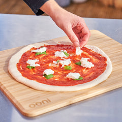 
A hand adds fresh mozzarella to a tomato and basil pizza being prepared on a bamboo peel engraved with the ooni logo.