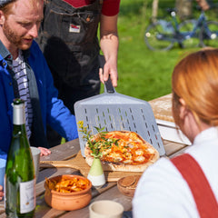 A person slides a cooked pizza onto a bamboo serving board using a perforated pizza peel.