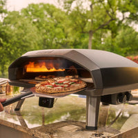 Ooni Koda 2 Max outdoor pizza oven cooking two pizzas, one already in the oven and another being inserted on a pizza peel in a backyard.