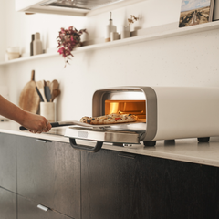 Person taking a pizza out of the White Ooni Volt 2 Pizza Oven in a kitchen.