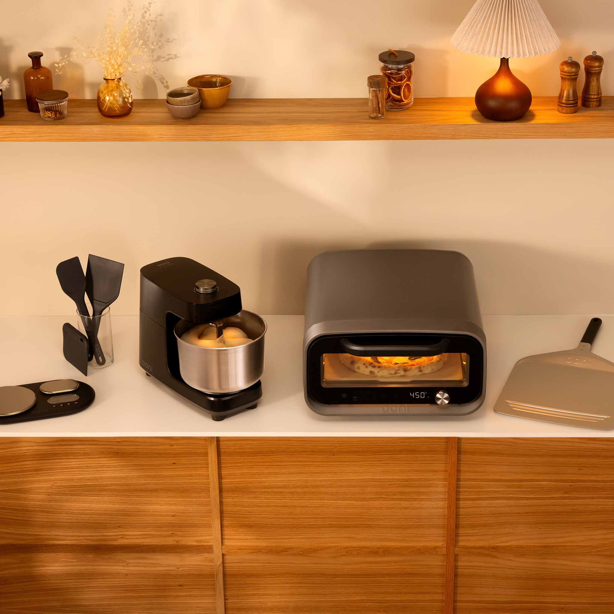A countertop kitchen scene showing a Volt 2 pizza oven with pizza inside, next to a Halo Core mixer with a stainless steel bowl, with a digital scale and a pizza peel on a light wood cabinet. A wooden shelf with decorative items is visible above.