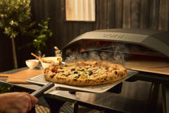 A steaming zucchini and olive pizza being pulled from an Ooni Koda 2 Max pizza oven during an outdoor evening cookout.