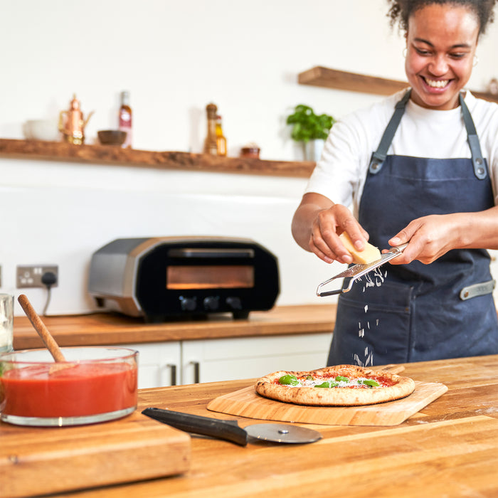Grating cheese on pizza with Ooni Volt 12 on kitchen counter | Click this image to open up the product gallery modal. The product gallery modal allows the images to be zoomed in on.